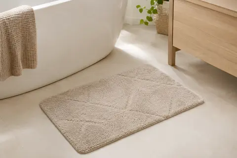 Tapis de bain rectangulaire beige à poil haut posé sur un sol clair dans une salle de bain lumineuse.