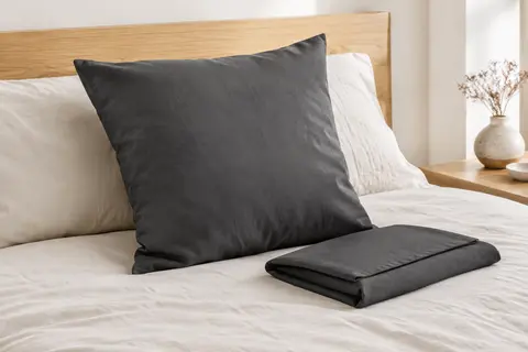 Deux taies d’oreiller anthracite en microfibre, l’une posée sur un lit crème et l’autre pliée.
