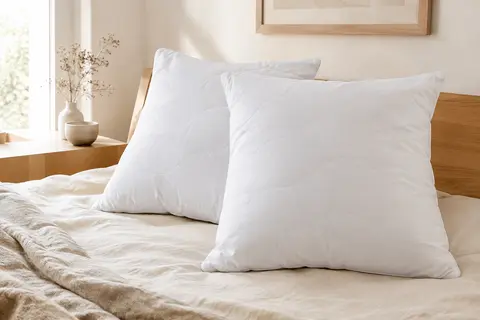 Deux oreillers carrés blancs à enveloppe matelassée posés sur un lit clair aux draps crème.