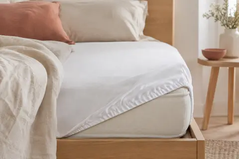 Protège-matelas blanc en jersey polyester installé sur un lit aux draps crème, dans une chambre lumineuse.