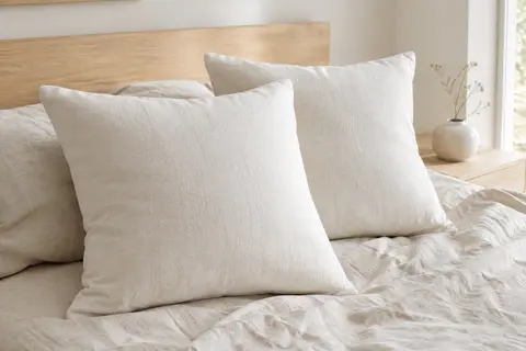 Deux coussins décoratifs carrés blancs posés sur un lit en lin froissé dans une chambre lumineuse.