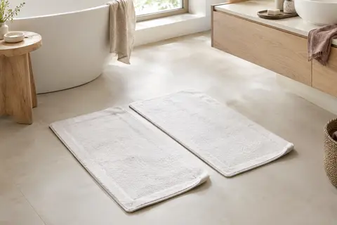Deux tapis de bain rectangulaires blancs en coton posés sur un sol clair dans une salle de bain lumineuse.